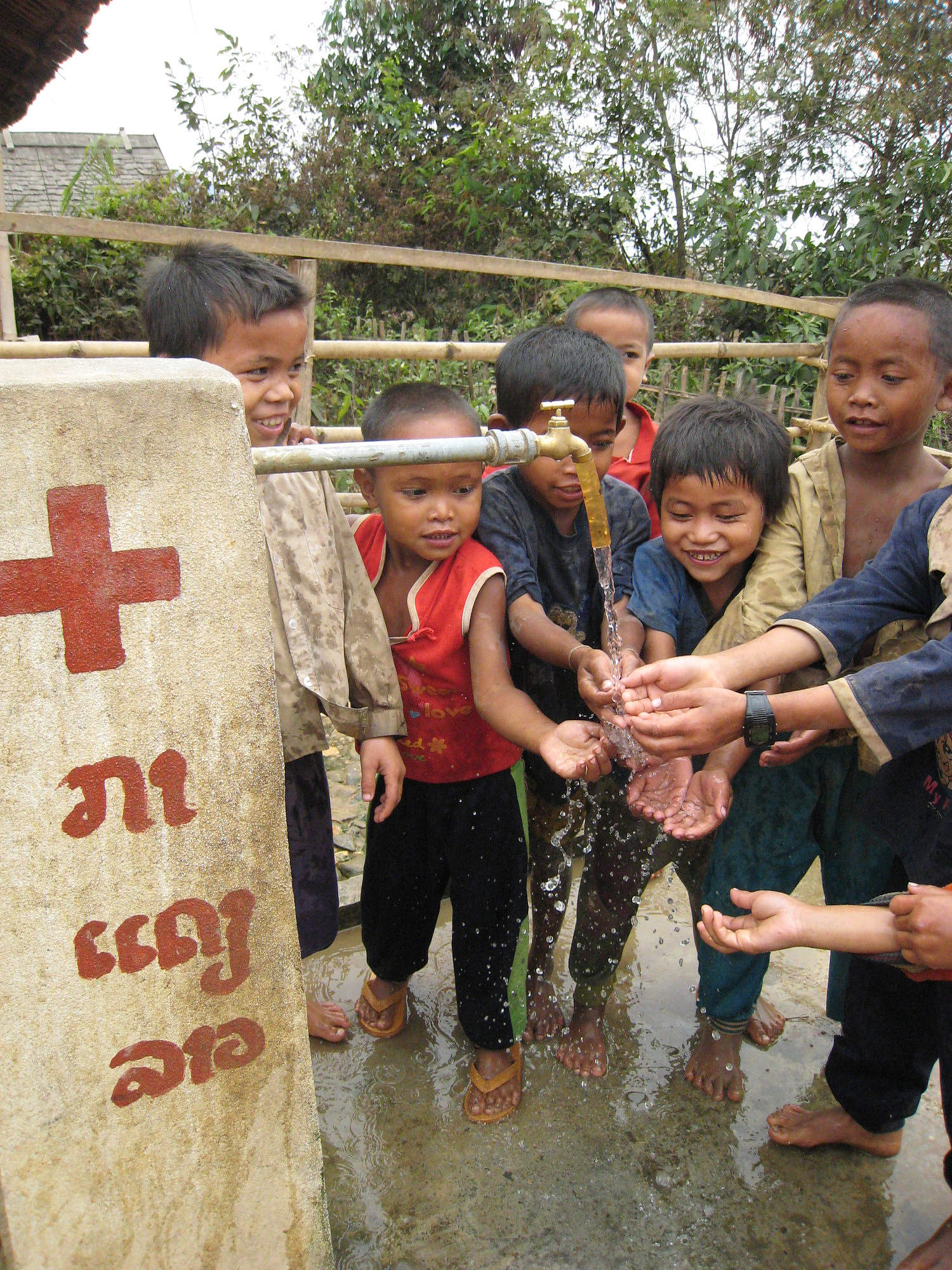 In Laos halten mehrere kleine Buben ihre Hände in das Wasser, das aus einem Brunnen kommt, und lachen.